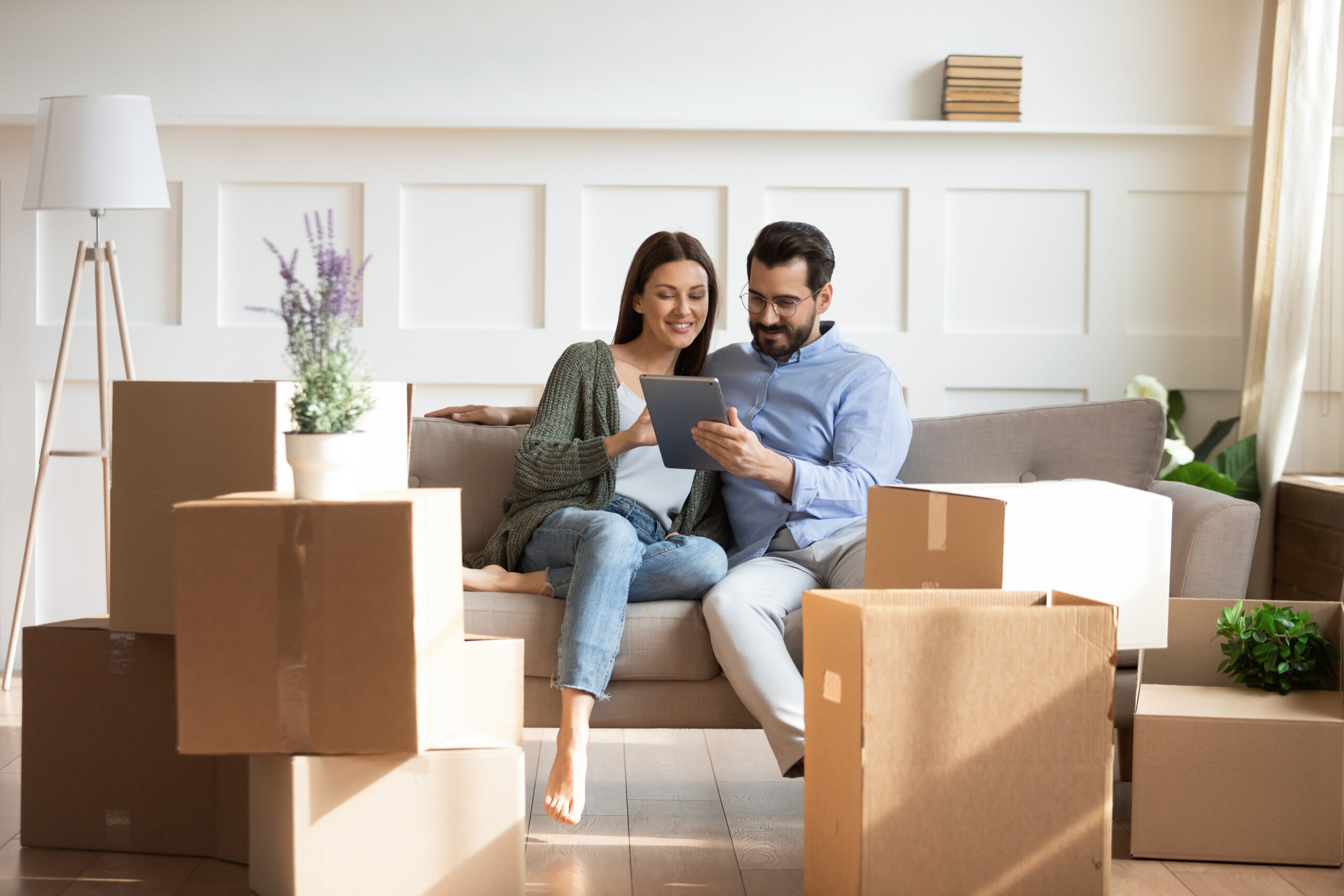 A couple sits on a couch surrounded by moving boxes, smiling as they look together at a tablet in their new home.