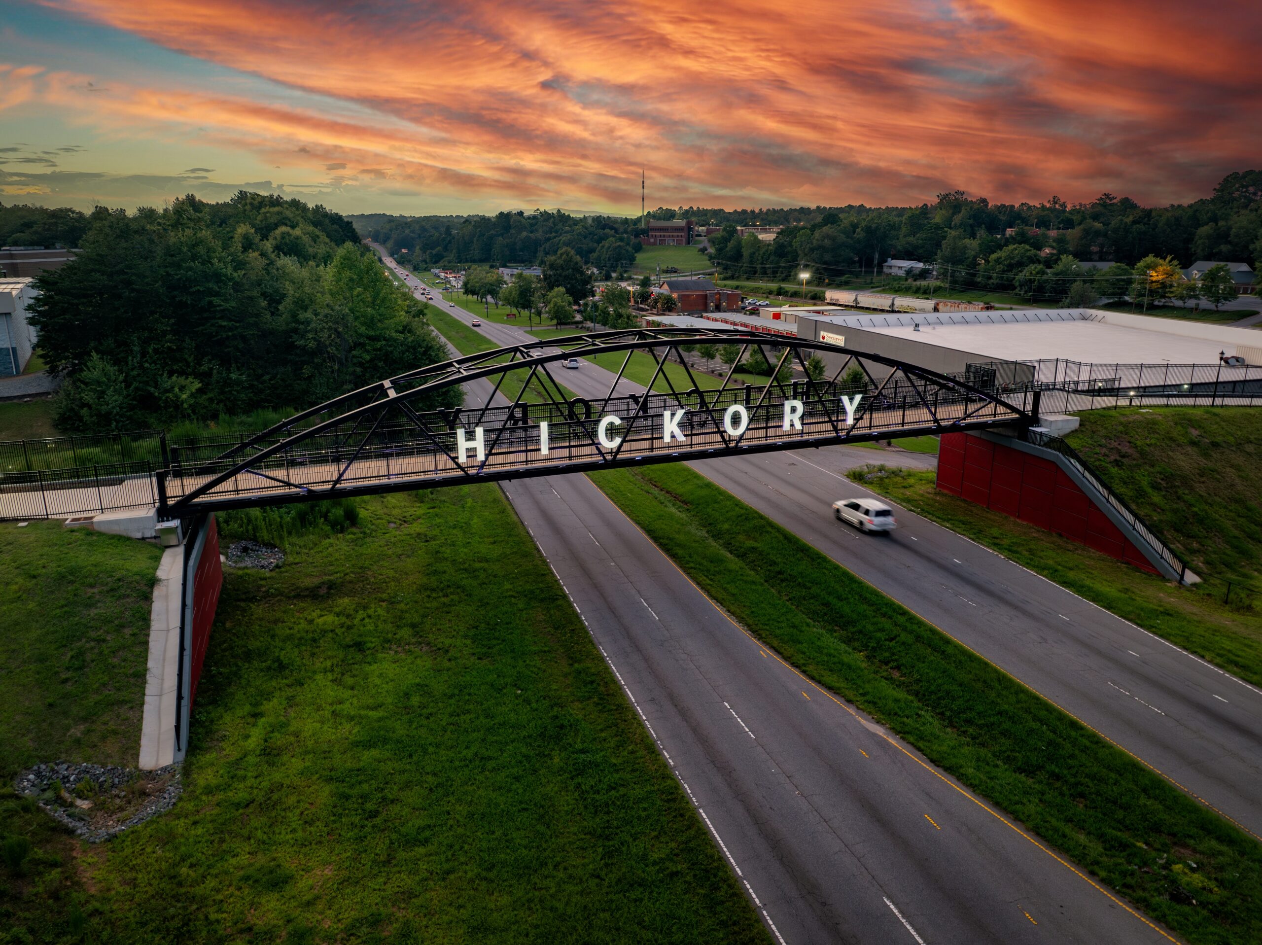 A pedestrian bridge with large white letters spelling “Hickory” spans over a highway at sunset, with cars driving below and trees and buildings in the background.